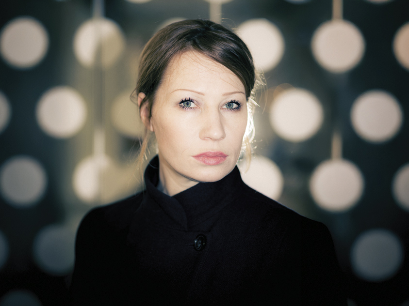Birgit Minichmayr, mit heller Haut und glattem braunem Haar, steht vor verschwommenen großen weißen, kreisförmigen Lichtern. Sie trägt einen dunklen Mantel und blickt mit neutraler Miene in die Kamera. 