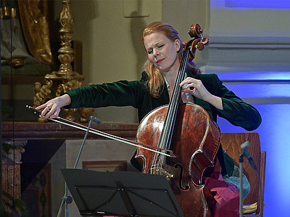 Harriet Krijgh sitzt auf einem Stuhl und spielt Cello beim beim Weihnachtskonzert 2025 der Salzkammergut Festwochen Gmunden in der festlich geschmückten Stadtpfarrkirche St. Nikolaus Bad Ischl. Sie trägt ein dunkelgrünes Samtoberteil und einen roten Rock. Das Cello ist braun und hat eine glänzende Oberfläche. Vor ihr steht ein Notenständer mit Notenblättern. Im Hintergrund sind eine goldene Kerzenhalterung und eine Wand mit blauer Beleuchtung zu sehen.