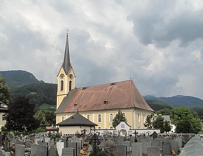 ​„Außenansicht der Evangelischen Kirche Bad Goisern: Ein klassizistisches Gebäude mit gelber Fassade, spitzem Kirchturm und umliegendem Friedhof