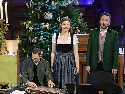 Die Musiker Rafael Fingerlos, Anja Mittermüller und Fabian Steindl bei einem Weihnachtskonzert 2025 er Salzkammergut Festwochen Gmunden vor einem geschmückten Christbaum, die zwei Männer in Anzug und Trachtenjacke, die Frau in Dirndl, Fabian Steindl spielt Zither.