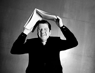 Ivor Bolton steht vor einer glatten Wand und hält ein geöffnetes Notenbuch über seinem Kopf, sodass die Seiten wie ein Dach wirken. Er trägt einen dunklen Anzug und lächelt direkt in die Kamera. Die Aufnahme ist schwarz-weiß und zeigt eine Nahaufnahme, die den Oberkörper und das Gesicht betont.