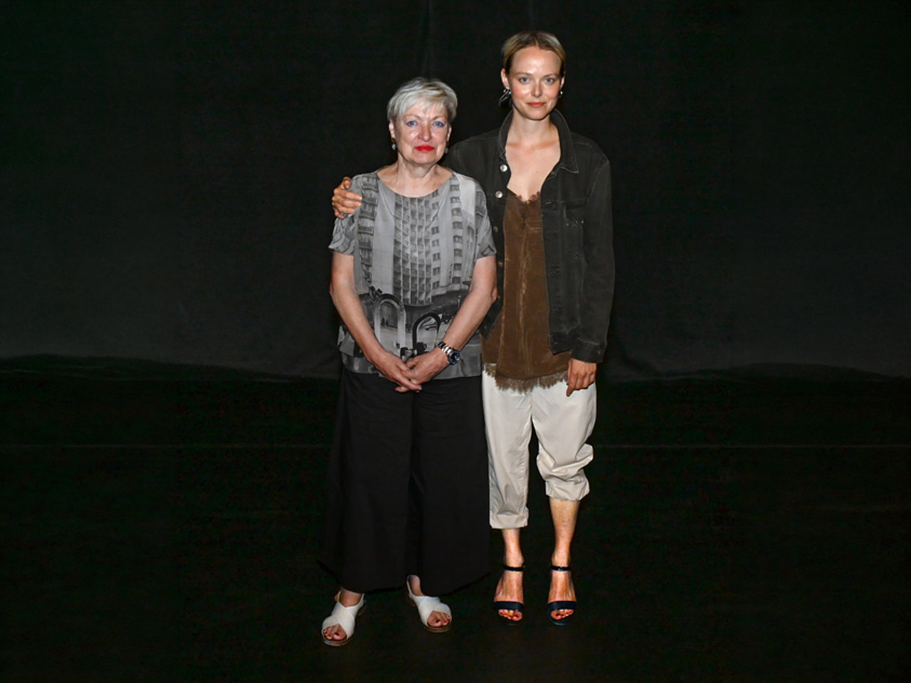 Karin Bergmann und Marie Luise Stockinger stehen nebeneinander vor dunklem Hintergrund. Karin Bergmann trägt eine helle gemusterte Bluse, eine dunkle weite Hose und weiße Sandalen. Marie Luise Stockinger trägt eine helle, locker sitzende Hose, ein braunes Oberteil und eine dunkle Jacke sowie schwarze Sandalen. Marie Luise Stockinger hat einen Arm um Karin Bergmann gelegt. Beide blicken in die Kamera.