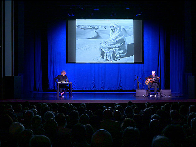 Christoph Ransmayr sitzt links am Tisch, Wolfgang Muthspiel rechts mit Gitarre auf der Bühne im Stadttheater Gmunden, Projektion eines Mannes in Wüstenkleidung, Salzkammergut Festwochen 25.05.2025