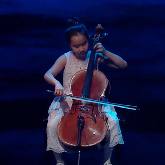 Ein junges Mädchen in einem weißen Kleid sitzt auf der Bühne und spielt Cello.