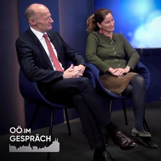 Zwei Personen sitzen nebeneinander auf dunkelblauen Stühlen in einem Studio. Der Text lautet: „OÖ IM GESPRÄCH“.