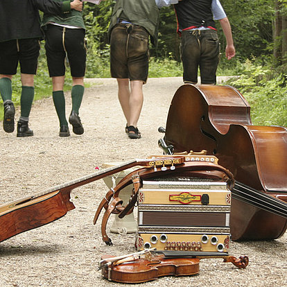 Drei Personen in traditioneller Kleidung gehen auf einem Waldweg davon, während im Vordergrund Musikinstrumente platziert sind.