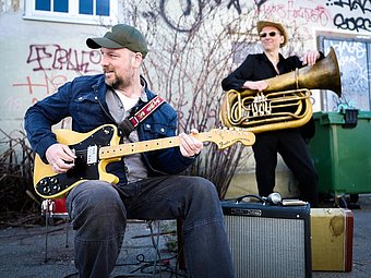 Stephan Zinner sitzt im Vordergrund auf einem Stuhl und spielt eine E-Gitarre. Er trägt eine blaue Jeansjacke, ein helles T-Shirt und eine Baseballkappe. Die Gitarre ist mit einem auffälligen Gurt versehen. Neben ihm steht ein Gitarrenverstärker. Im Hintergrund steht eine weitere Person mit einer Tuba, beide befinden sich im Freien vor einer Wand mit Graffiti und kahlen Ästen.