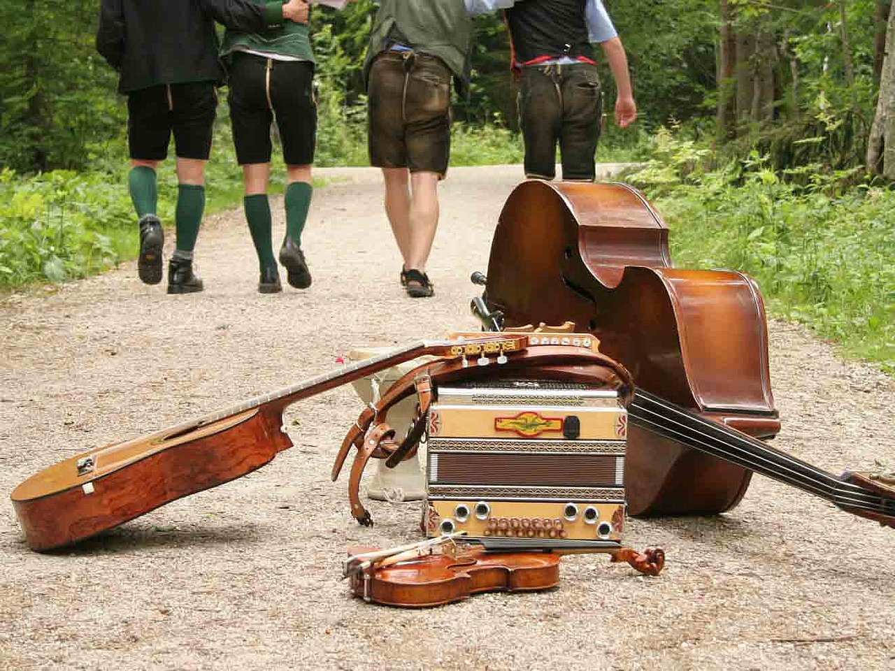 Mehrere Musikinstrumente, darunter ein Akkordeon, eine Geige, eine Gitarre und ein Kontrabass, liegen auf einem Waldweg, während im Hintergrund drei Personen in traditioneller Kleidung weggehen.