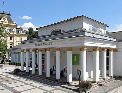 "Außenansicht der klassizistischen Trinkhalle in Bad Ischl mit weißer Fassade, goldverzierten korinthischen Säulen