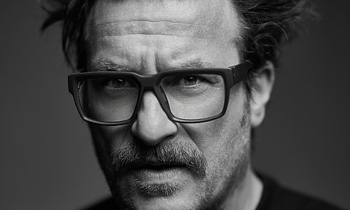 Marcus Füreder, bekannt als Parov Stelar, trägt ein dunkles Oberteil. Das Bild ist in schwarz-weiß gehalten. Sein Haar ist mittellang und wirkt leicht zerzaust, mit einzelnen Strähnen, die nach oben und zur Seite stehen. Der Hintergrund ist einfarbig und neutral, ohne erkennbare Details.