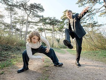 Zwei Mitglieder von Wiener Blond posieren auf einem Waldweg in ausdrucksstarker Haltung, beide tragen elegante Kleidung und schauen direkt in die Kamera.