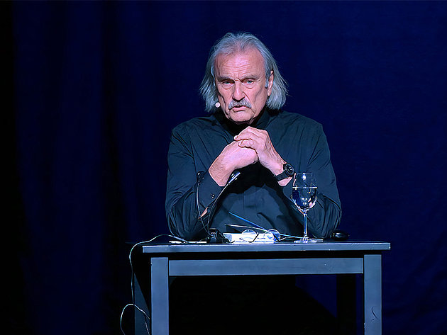 Christoph Ransmayr sitzt bei einer Lesung im Stadttheater Gmunden an einem Tisch mit einem Glas Wasser und Mikrofon