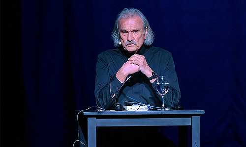 Christoph Ransmayr sitzt bei einer Lesung im Stadttheater Gmunden an einem Tisch mit einem Glas Wasser und Mikrofon