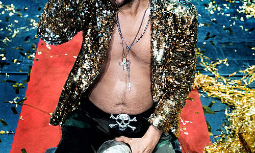 Philipp Hochmair kniet auf einem roten Teppich mit offenem Hemd, Glitzerjacke, Camouflagehose und hält einen Totenschädel in der Hand, umgeben von goldenen Konfetti.